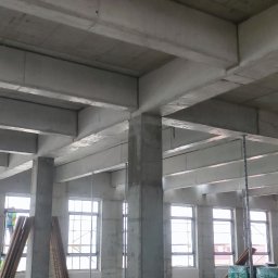 VibeTom - Surowy szkielet budynku żelbetowego w trakcie budowy. Widoczne betonowe słupy, stropy i ściany z oknami. Elementy szalunków i podpór. W tle widoczny pracownik.