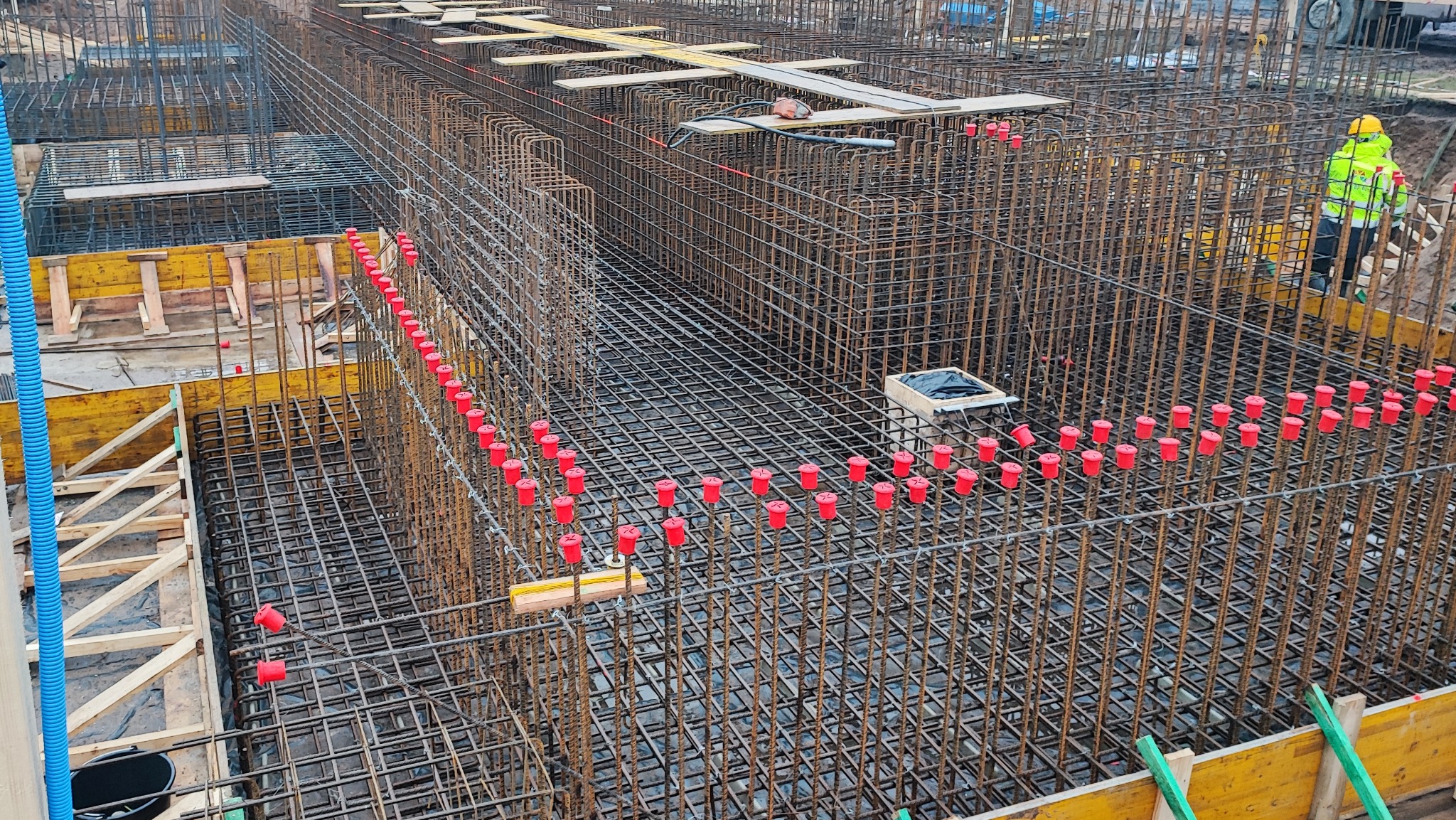 Skomplikowane zbrojenie fundamentów z czerwonymi osłonami na prętach, widoczne szalunki i pracownik w odblaskowej kurtce w tle. Detal budowy.