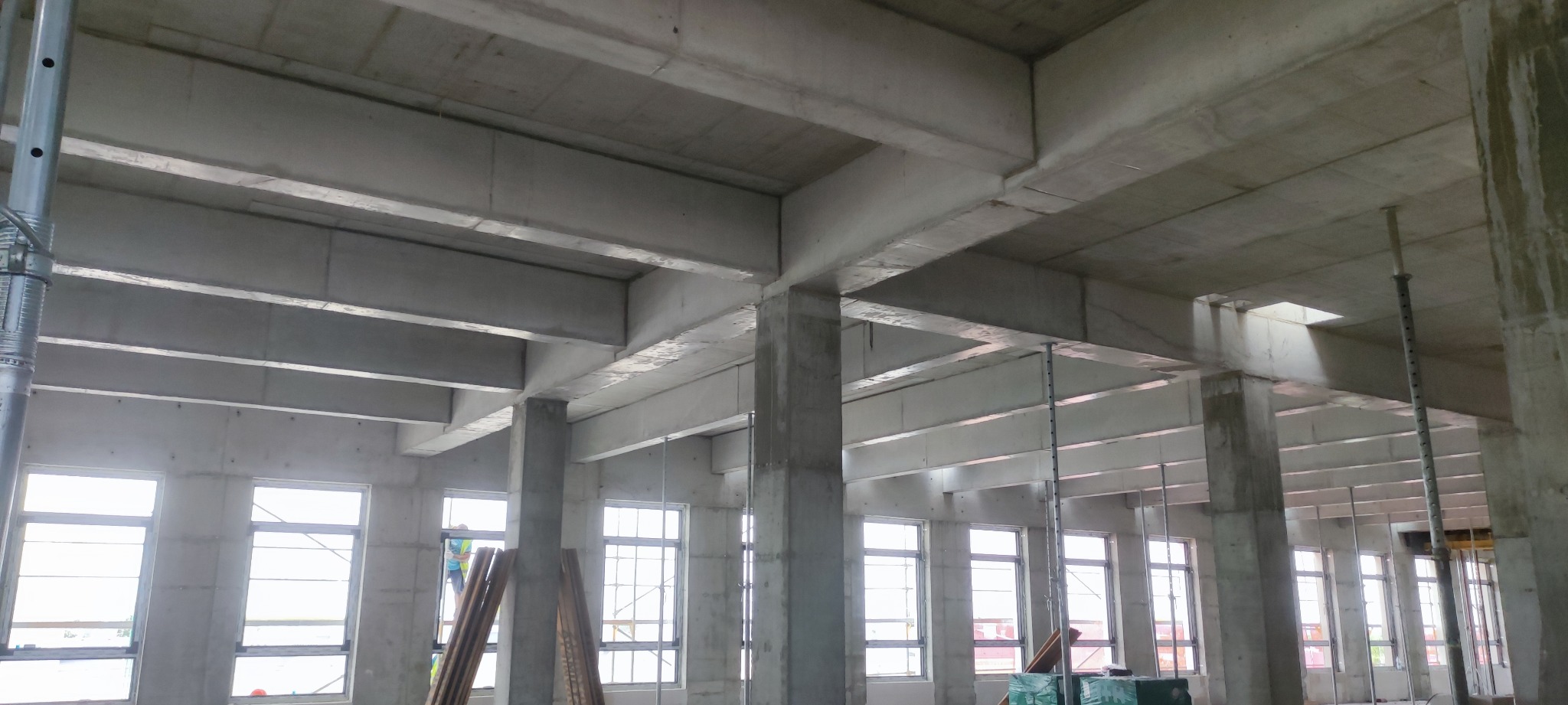 Surowy szkielet budynku żelbetowego w trakcie budowy. Widoczne betonowe słupy, stropy i ściany z oknami. Elementy szalunków i podpór. W tle widoczny pracownik.