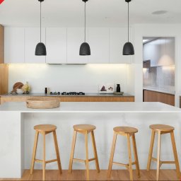 SOFIX KUCHNIE - Jasna kuchnia z drewnianymi akcentami, wyspą z białym blatem i czterema drewnianymi hokerami, oświetlona trzema wiszącymi lampami z czarnymi abażurami, widoczne schody i rośliny w wazonie.