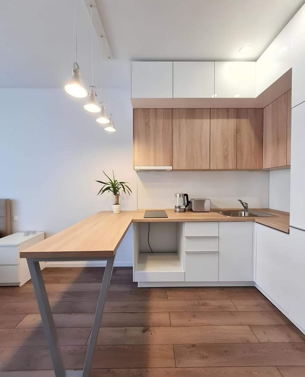 Nowoczesna kuchnia z drewnianymi szafkami wiszącymi, białymi szafkami dolnymi i blatem z przedłużonym półwyspem wspartym na metalowej konstrukcji w kształcie litery V. Na blacie czajnik, toster...