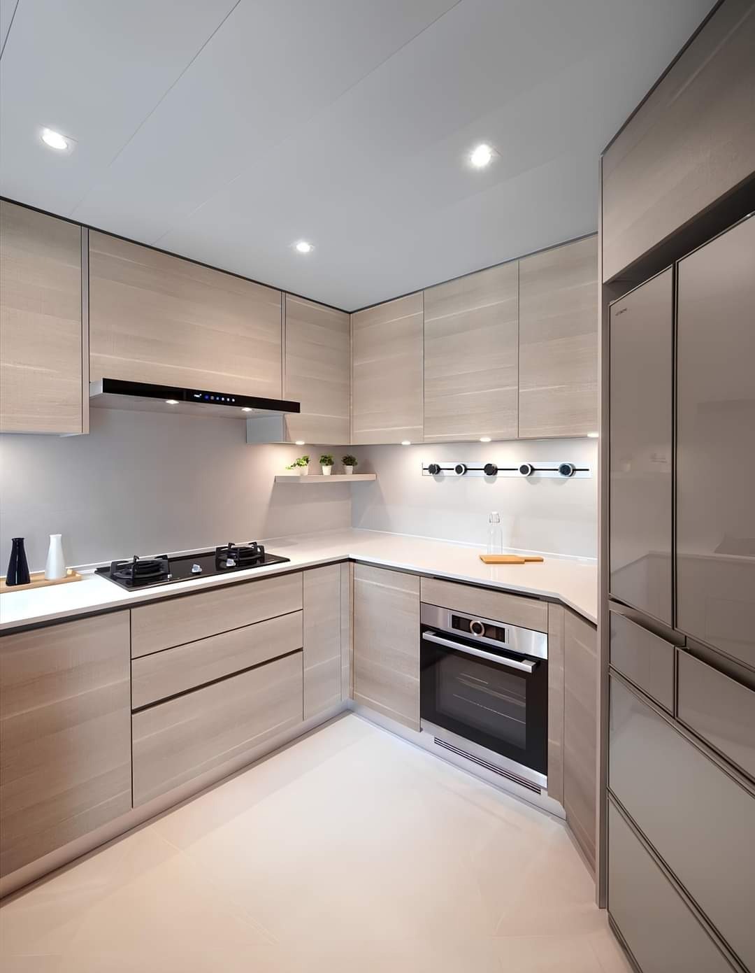 Nowoczesna kuchnia w jasnych odcieniach drewna, z minimalistycznym oświetleniem punktowym, czarną płytą grzewczą i piekarnikiem w zabudowie, uzupełniona o dekoracyjne flakoniki i małe rośliny.