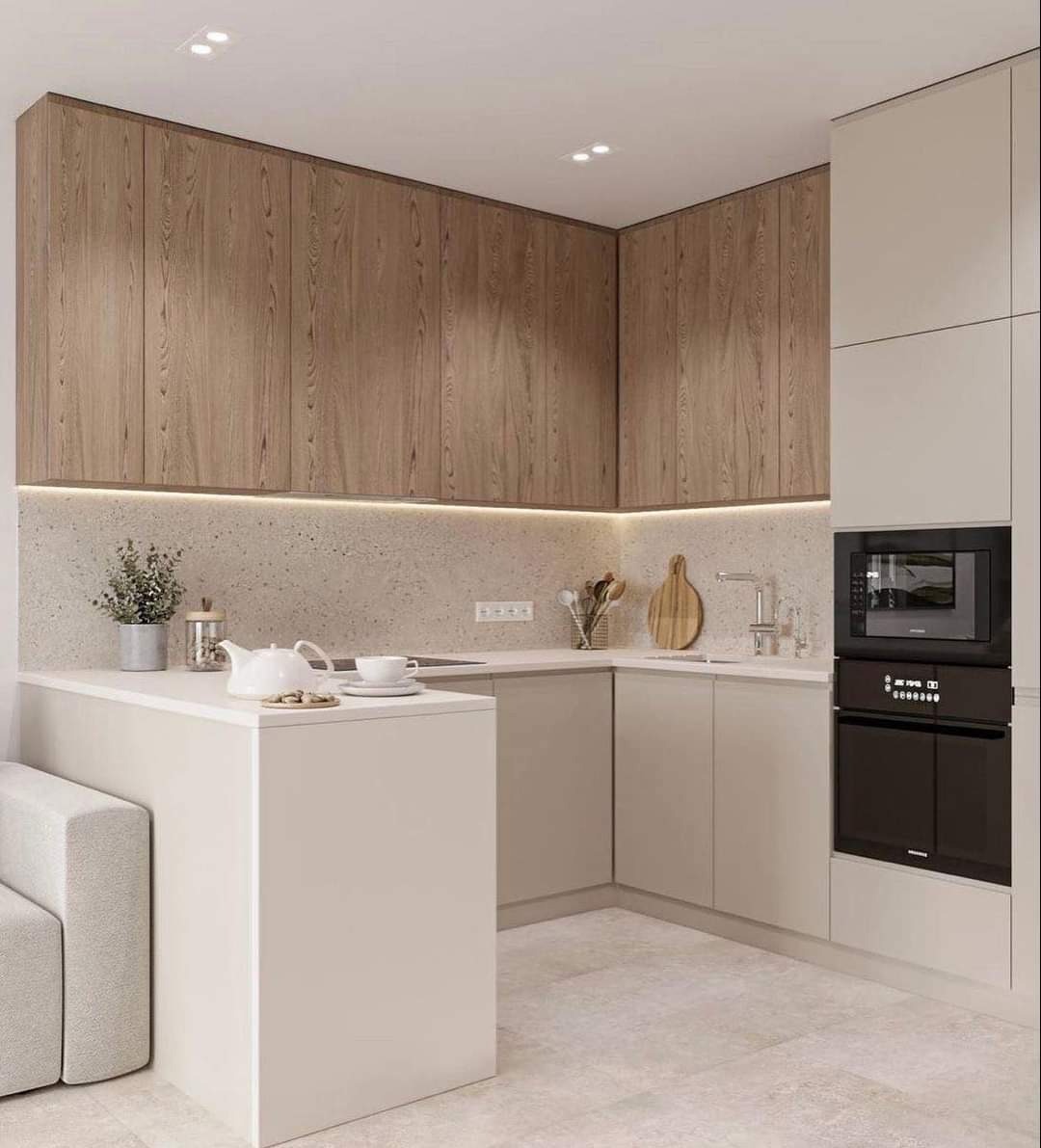 Jasna kuchnia w stylu minimalistycznym z drewnianymi szafkami górnymi, oświetleniem LED pod szafkami i wyspą z białym blatem, na której stoi czajnik, filiżanka i roślina doniczkowa.