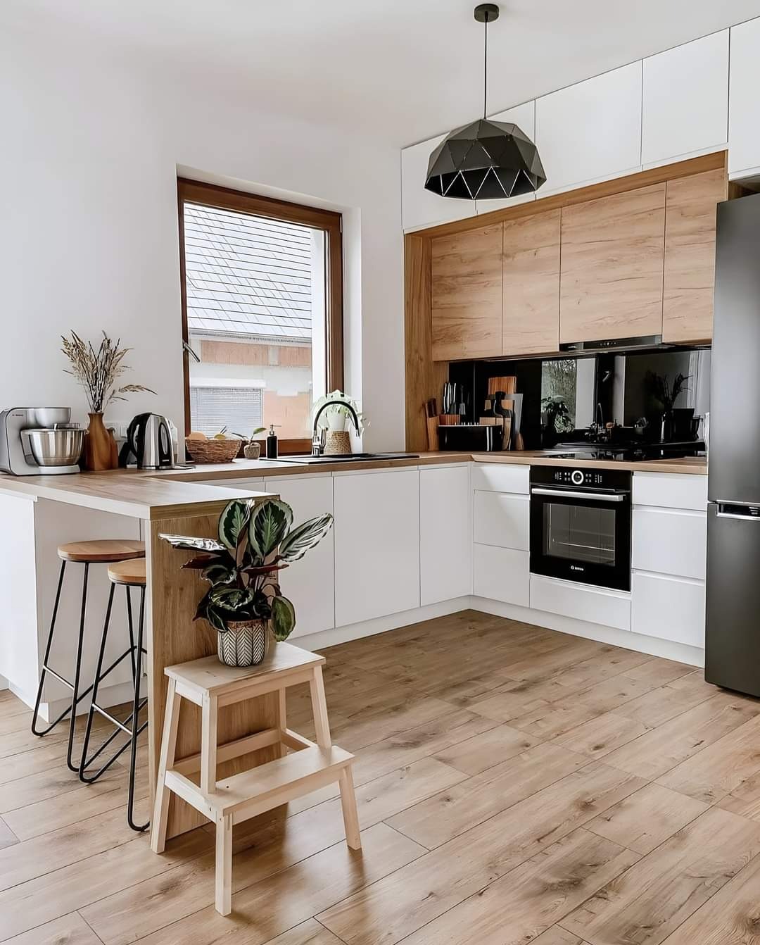 Jasna kuchnia w stylu skandynawskim z drewnianymi akcentami: białe szafki, drewniany blat, czarny piekarnik i lodówka, drewniana podłoga, roślina doniczkowa na drewnianym stołku, nowoczesna lampa...