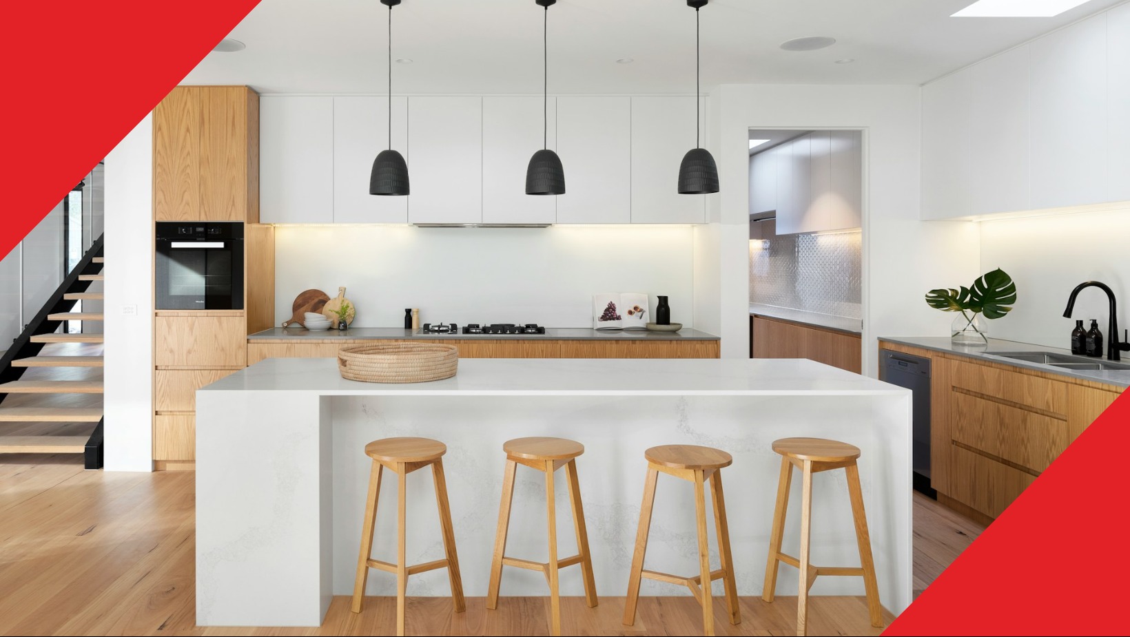 Jasna kuchnia z drewnianymi akcentami, wyspą z białym blatem i czterema drewnianymi hokerami, oświetlona trzema wiszącymi lampami z czarnymi abażurami, widoczne schody i rośliny w wazonie.