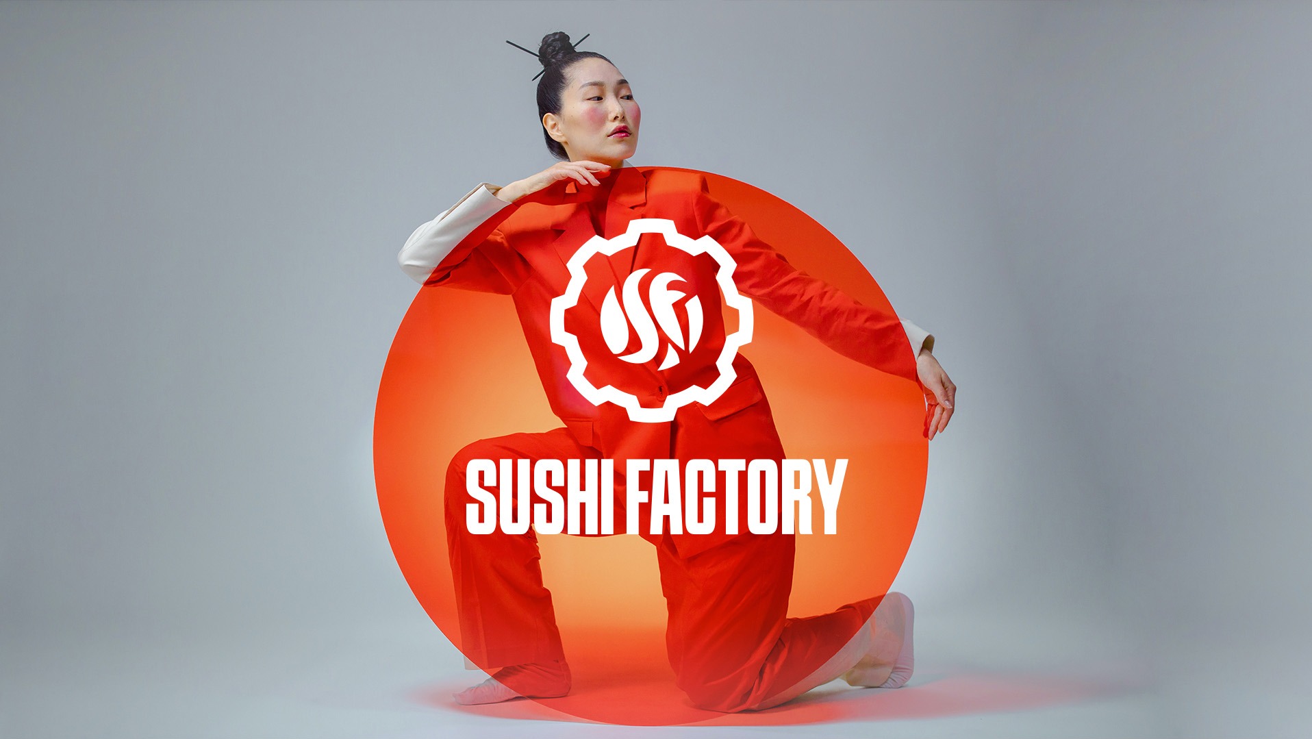 Kobieta w czerwonym garniturze pozuje na kolanach za okręgiem z logo i napisem Sushi Factory na szarym tle.