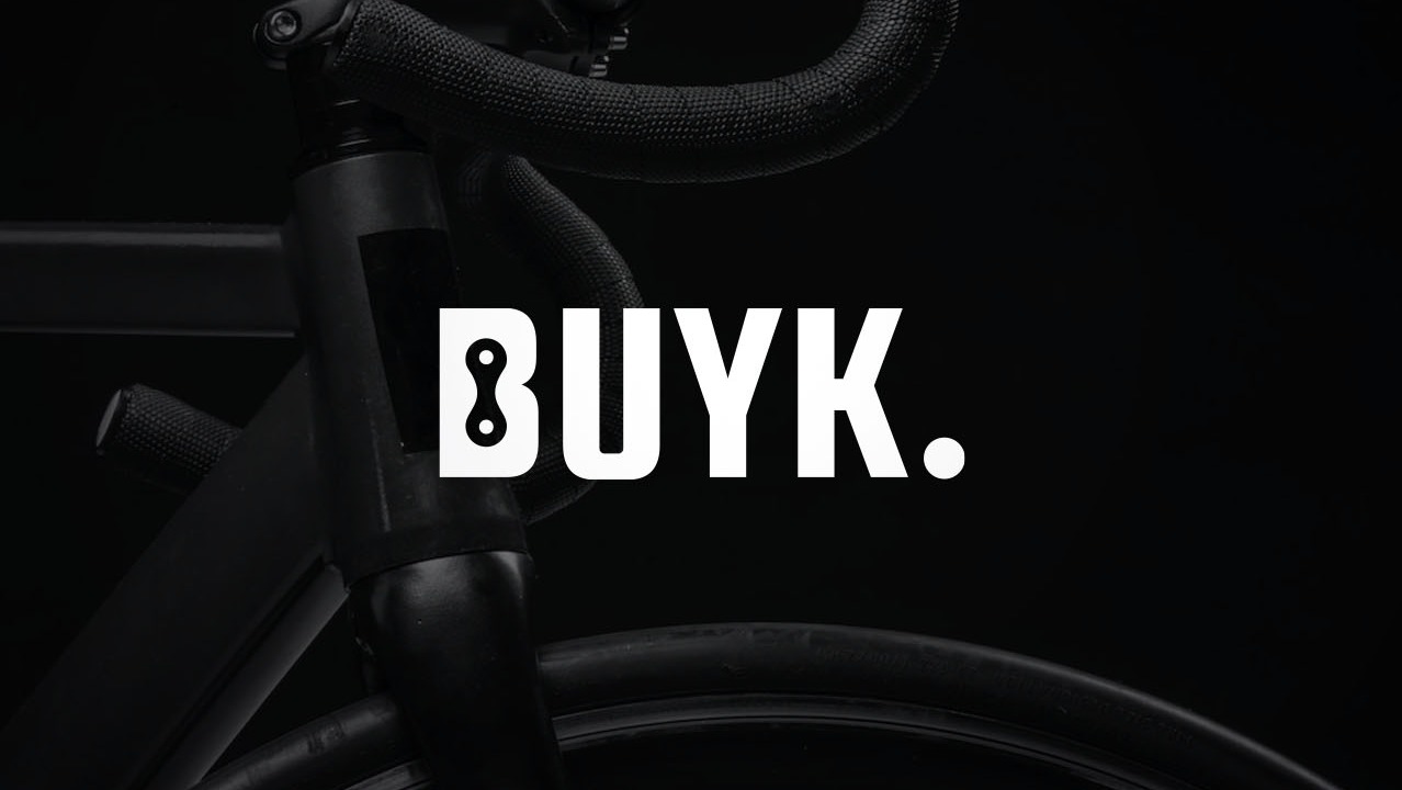 Minimalistyczne ujęcie roweru z widocznym logo 'BUYK' na czarnym tle, prezentujące detale kierownicy i opony.