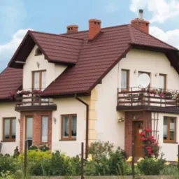 Dwupiętrowy dom z ciemnoczerwoną dachówką, balkonem z kwiatami i anteną satelitarną, otoczony zielenią i ogrodzeniem.