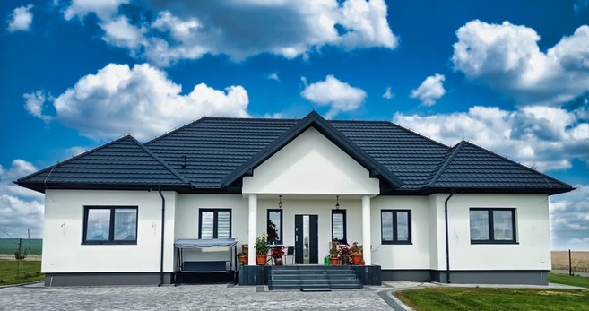 Nowoczesny dom jednorodzinny z ciemnoszarym dachem z dachówki ceramicznej, białymi ścianami i ciemnymi oknami, na tle błękitnego nieba z obłokami.