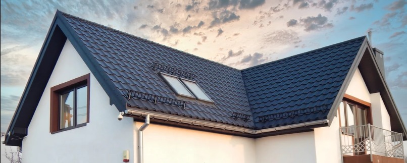 Dom jednorodzinny z ciemnym dachem z blachodachówki, dwoma oknami połaciowymi i orynnowaniem w kolorze srebrnym, na tle pochmurnego nieba.