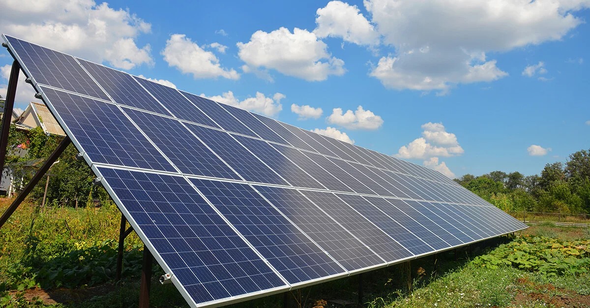 Panel słoneczny ustawiony pod kątem na metalowej konstrukcji, z widocznym ogrodem i domem w tle, na tle błękitnego nieba z białymi obłokami.