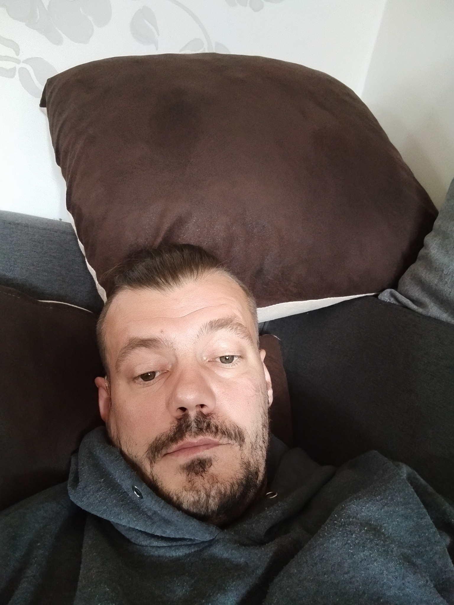 Mężczyzna z brodą leżący na szarej sofie z brązową poduszką na tle białej ściany z kwiatowym wzorem.