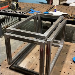 Marek Szmigiel - Spawana, przestrzenna konstrukcja z metalowych profili na perforowanym stole spawalniczym, w tle widoczny hełm spawalniczy i narzędzia. Surowy stan przed obróbką.