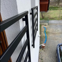 Marek Szmigiel - Dwa czarne, metalowe kwietniki zamontowane na elewacji domu z widocznymi mocowaniami, z perspektywą na ogród i podjazd.