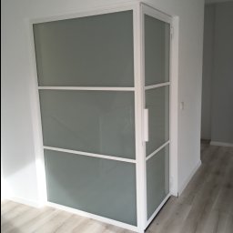 Marek Szmigiel - Nowoczesne drzwi wewnętrzne z matowego szkła i białej ramy, widoczne z boku, z uchwytem, na tle białych ścian i podłogi z paneli.