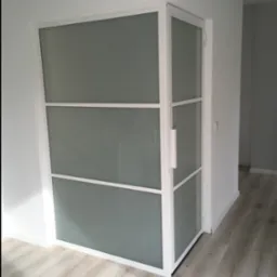 Nowoczesne drzwi wewnętrzne z matowego szkła i białej ramy, widoczne z boku, z uchwytem, na tle białych ścian i podłogi z paneli.