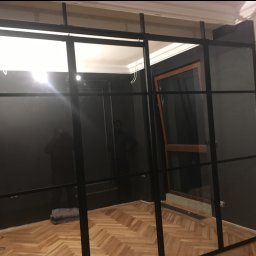 Marek Szmigiel - Nowoczesna, czarna konstrukcja stalowa z przeszkleniami w jasnym wnętrzu. Widoczny parkiet, ściany w dwóch kolorach, okno z drewnianą ramą. Częściowo widoczny fotograf.