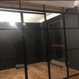 Nowoczesna, czarna konstrukcja stalowa z przeszkleniami w jasnym wnętrzu. Widoczny parkiet, ściany w dwóch kolorach, okno z drewnianą ramą. Częściowo widoczny fotograf.