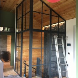 Marek Szmigiel - Nowoczesna, przeszklona konstrukcja stalowa wewnątrz domu z drewnianym wykończeniem, widoczna drabina i zabezpieczenia podłogi, sugerujące etap montażu.