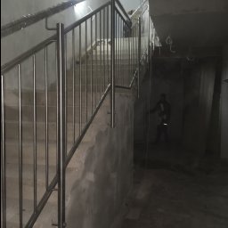 Marek Szmigiel - Nowa balustrada ze stali nierdzewnej, częściowo owinięta folią ochronną, zamontowana na betonowych schodach w surowym wnętrzu budynku w trakcie wykończenia.