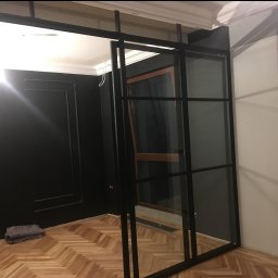 Marek Szmigiel - Loftowa, czarna, przeszklona konstrukcja działowa z metalu, oddzielająca pokoje. Widoczny parkiet i fragment czarnej ściany z dekoracyjnymi listwami.