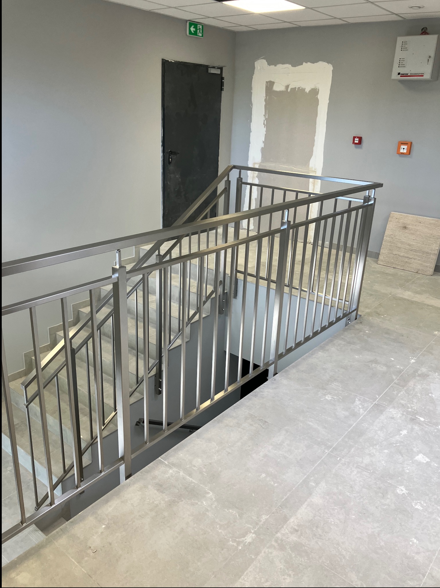 Nowoczesne, metalowe poręcze schodowe w szarym wnętrzu budynku. Widoczne płytki podłogowe, drzwi ewakuacyjne i fragment ściany z instalacją elektryczną.
