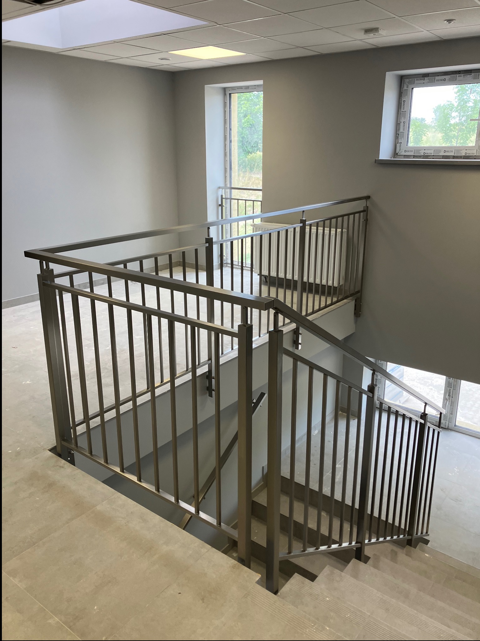 Metalowa poręcz schodowa w nowoczesnym budynku, widok z góry, szare ściany, naturalne światło z okien. Minimalistyczny design.