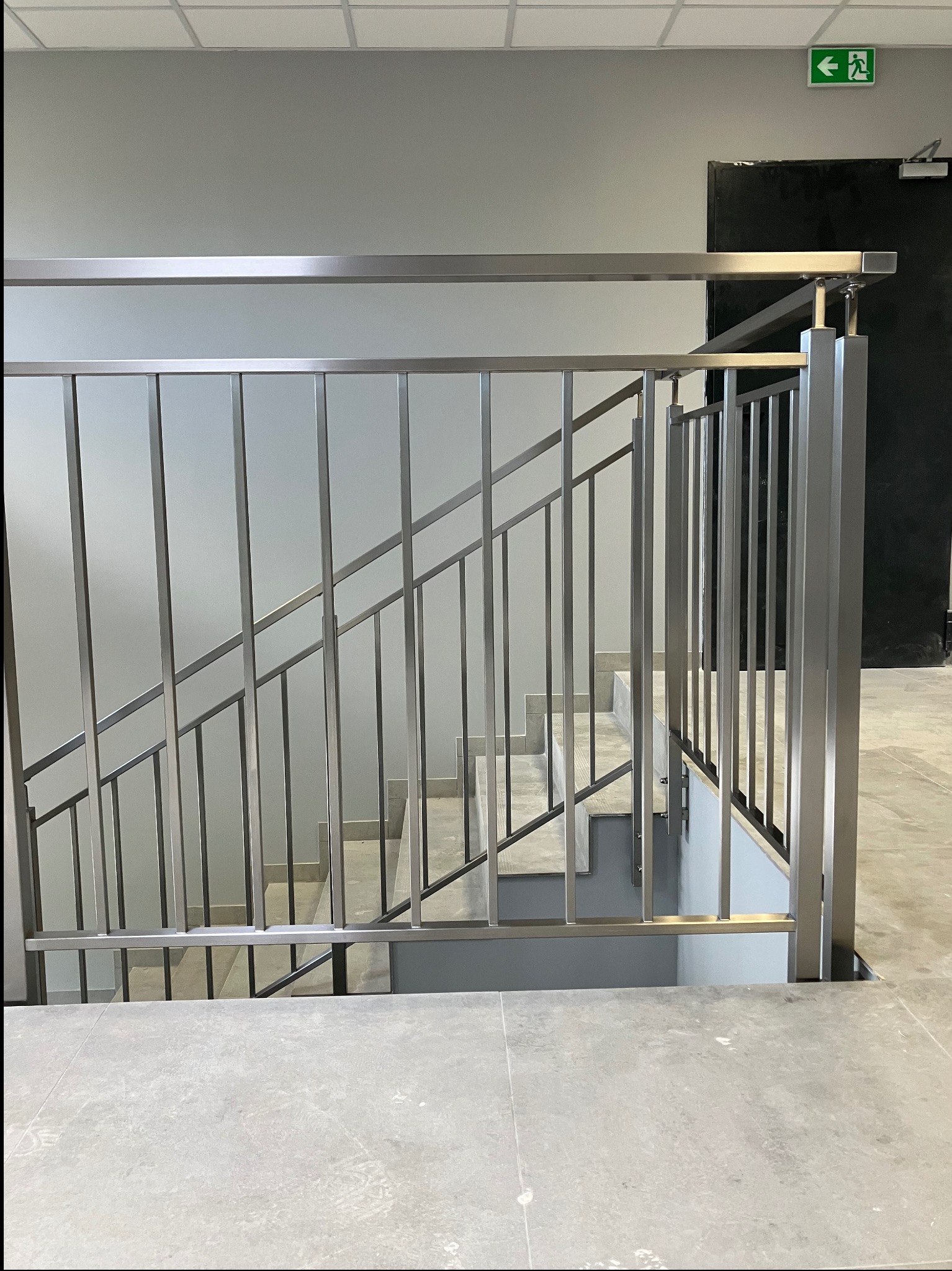 Wnętrze budynku z nowoczesnymi, metalowymi poręczami schodowymi o minimalistycznym designie. Widoczny znak ewakuacyjny nad drzwiami i szare płytki podłogowe.