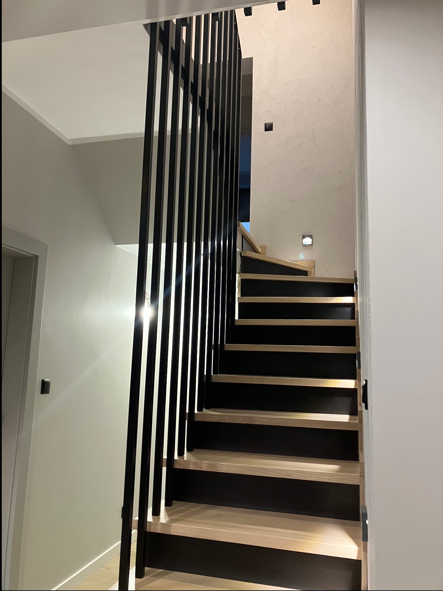 Nowoczesne schody z drewnianymi stopniami i pionową, czarną balustradą z metalowych prętów. Minimalistyczny design, jasne wnętrze, oświetlenie punktowe.