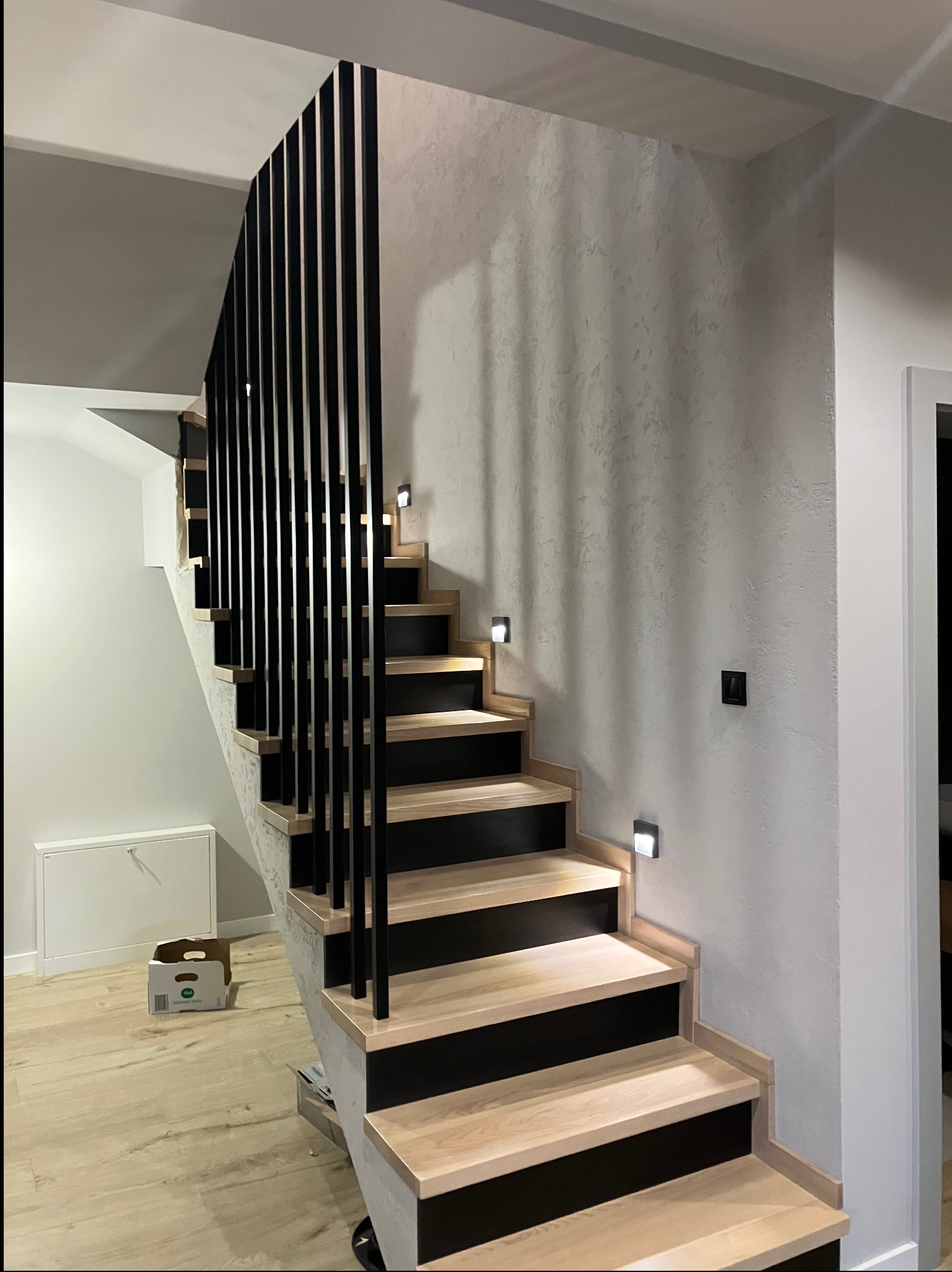 Nowoczesne schody z drewnianymi stopniami, czarnymi podstopnicami i minimalistyczną, pionową balustradą. Oświetlenie LED w ścianie, surowa faktura ściany.
