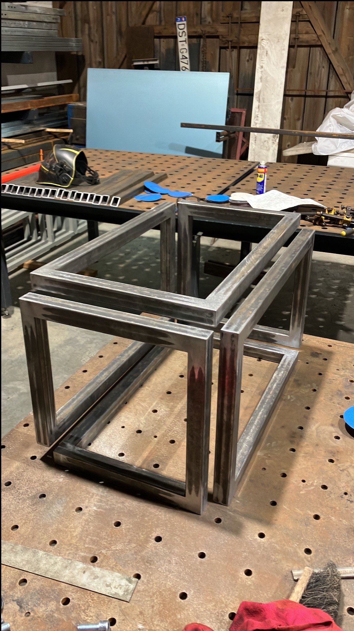 Spawana, przestrzenna konstrukcja z metalowych profili na perforowanym stole spawalniczym, w tle widoczny hełm spawalniczy i narzędzia. Surowy stan przed obróbką.