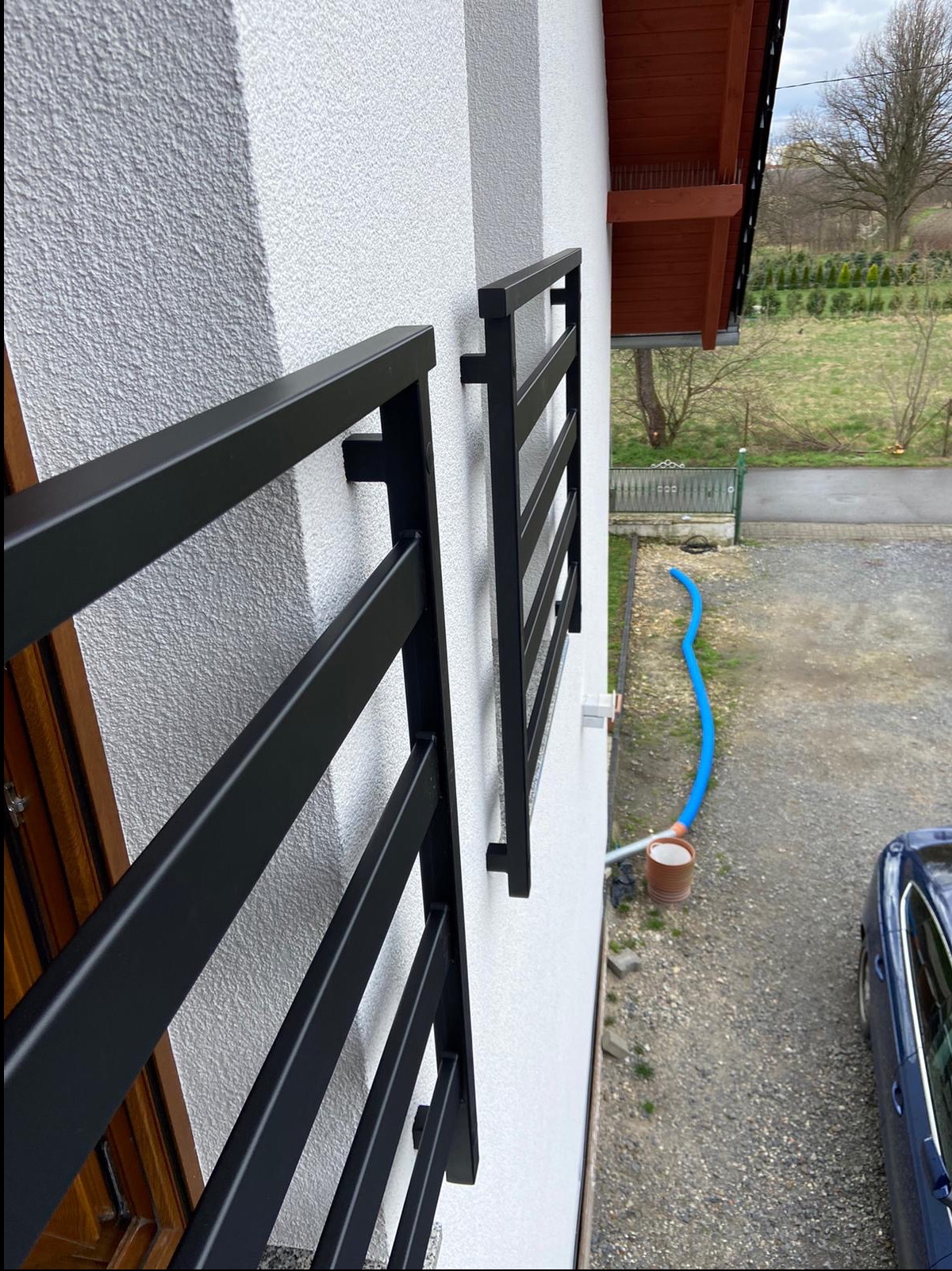 Dwa czarne, metalowe kwietniki zamontowane na elewacji domu z widocznymi mocowaniami, z perspektywą na ogród i podjazd.