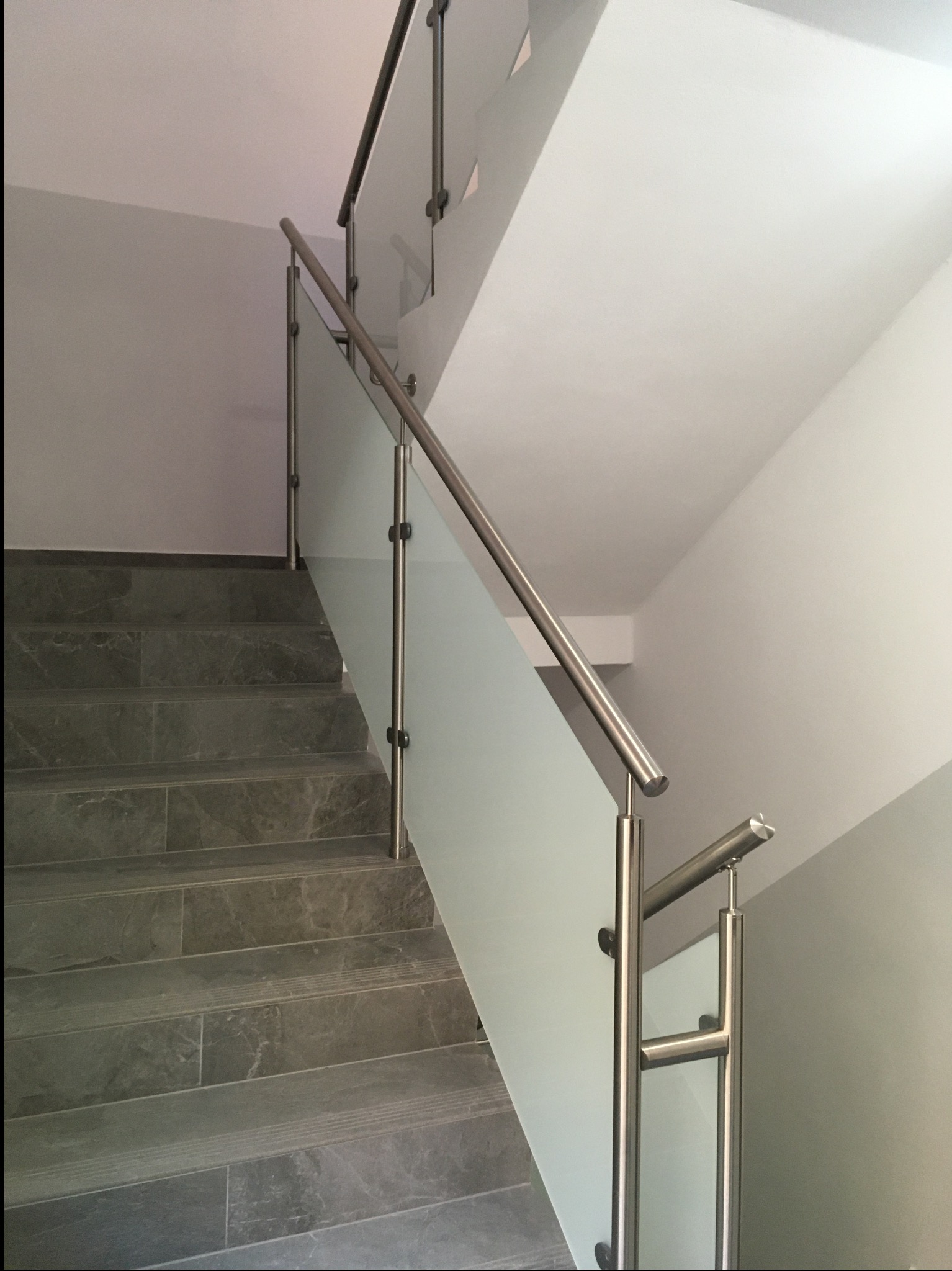 Szklana balustrada ze stali nierdzewnej zamocowana na schodach z szarych płytek. Widok z dołu, na pierwszym planie fragment poręczy.