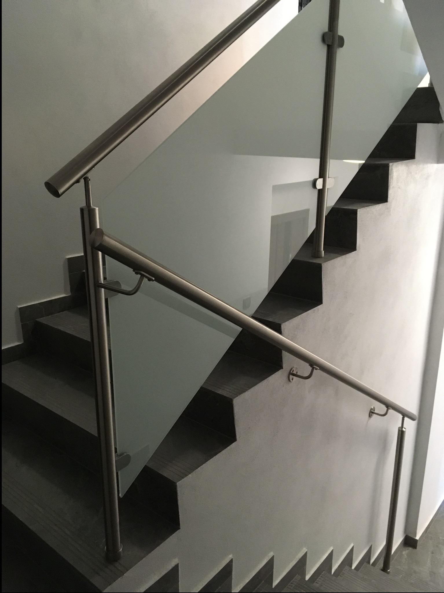 Szklana balustrada schodowa z nierdzewnymi poręczami, zamontowana wewnątrz budynku. Widok z góry, na schody z ciemnymi stopniami i jasnymi ścianami.