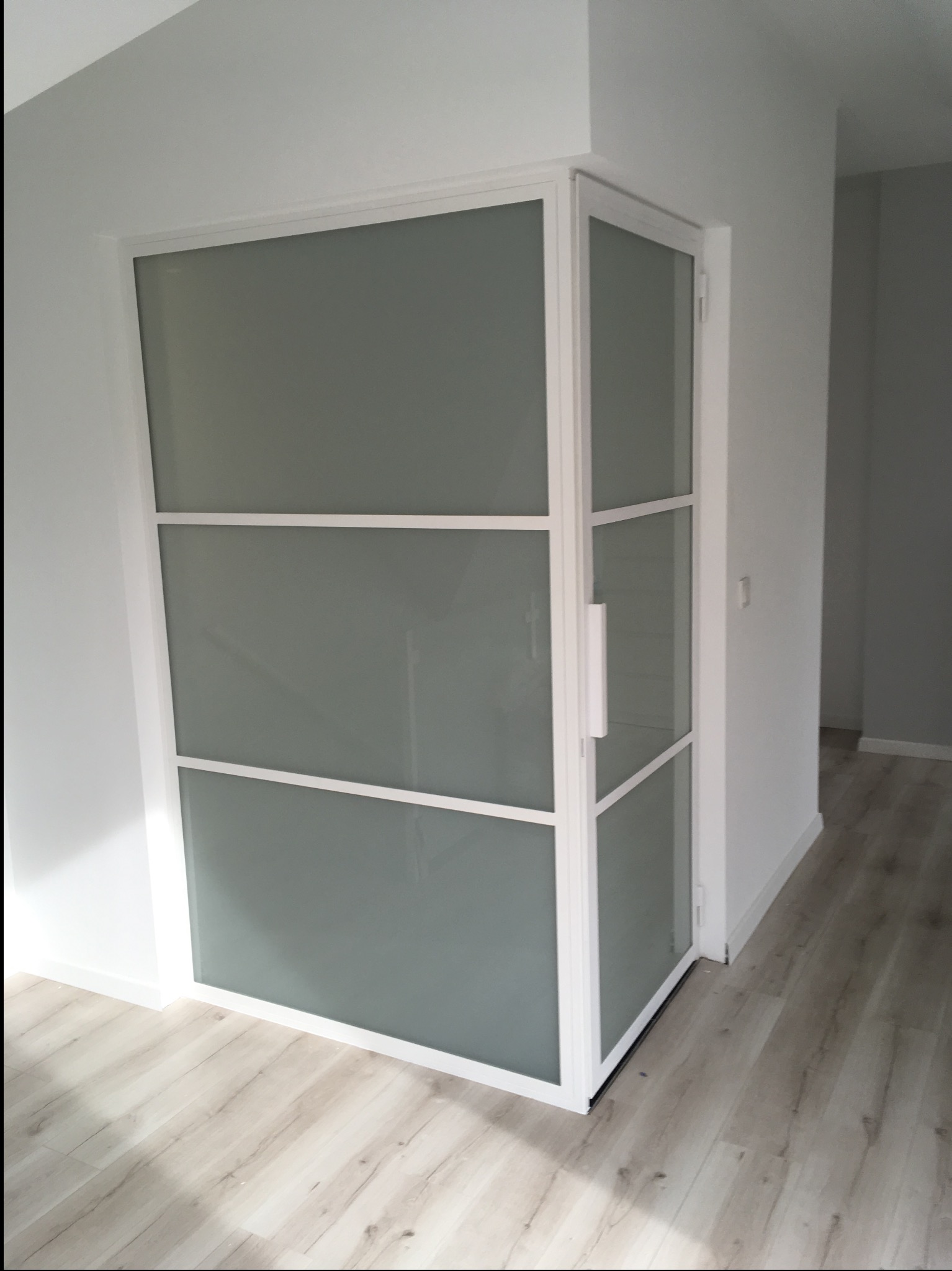 Nowoczesne drzwi wewnętrzne z matowego szkła i białej ramy, widoczne z boku, z uchwytem, na tle białych ścian i podłogi z paneli.