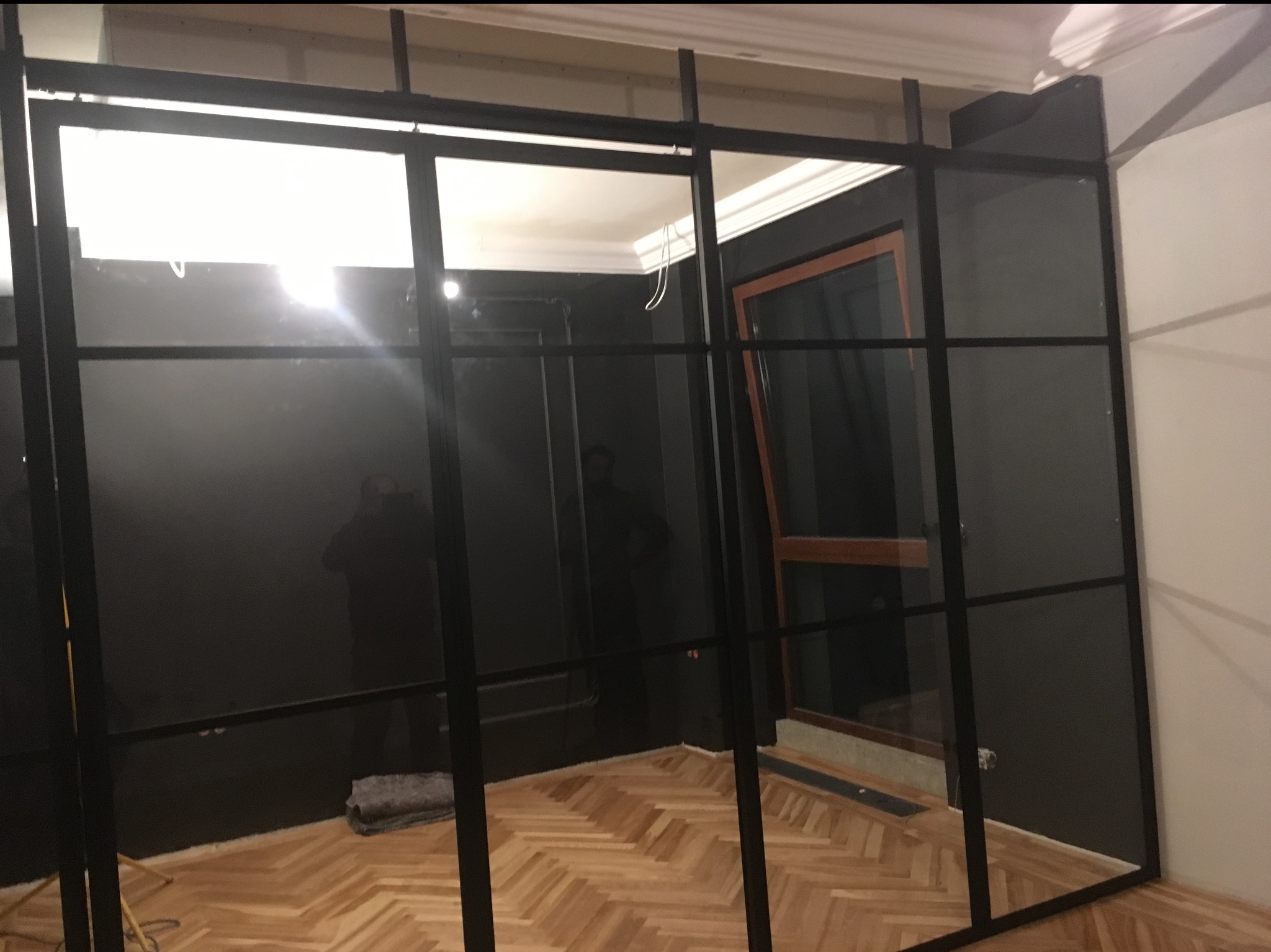 Nowoczesna, czarna konstrukcja stalowa z przeszkleniami w jasnym wnętrzu. Widoczny parkiet, ściany w dwóch kolorach, okno z drewnianą ramą. Częściowo widoczny fotograf.