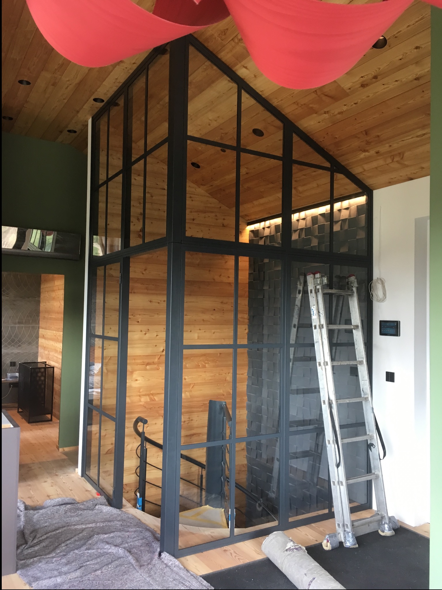 Nowoczesna, przeszklona konstrukcja stalowa wewnątrz domu z drewnianym wykończeniem, widoczna drabina i zabezpieczenia podłogi, sugerujące etap montażu.