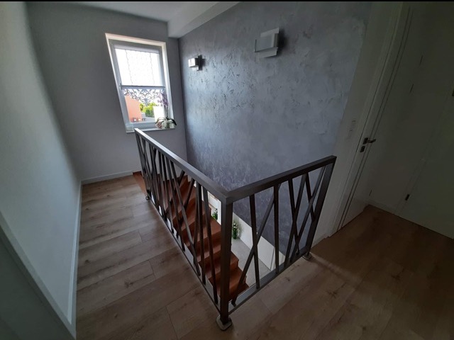 Metalowa poręcz schodowa z geometrycznym wzorem w nowoczesnym wnętrzu. Widok z góry na schody z drewnianymi stopniami i szarą ścianą z dekoracyjną fakturą.