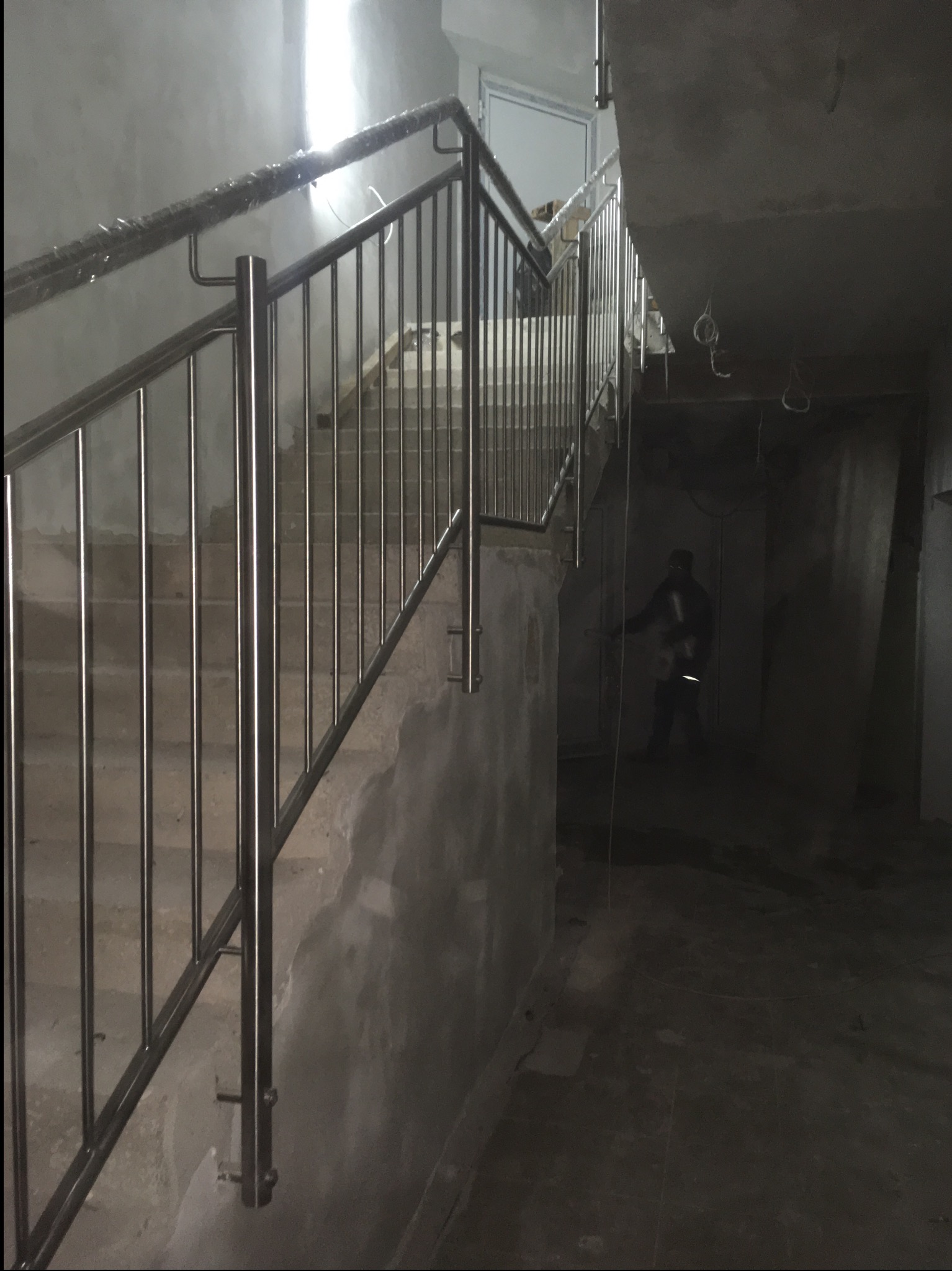 Nowa balustrada ze stali nierdzewnej, częściowo owinięta folią ochronną, zamontowana na betonowych schodach w surowym wnętrzu budynku w trakcie wykończenia.