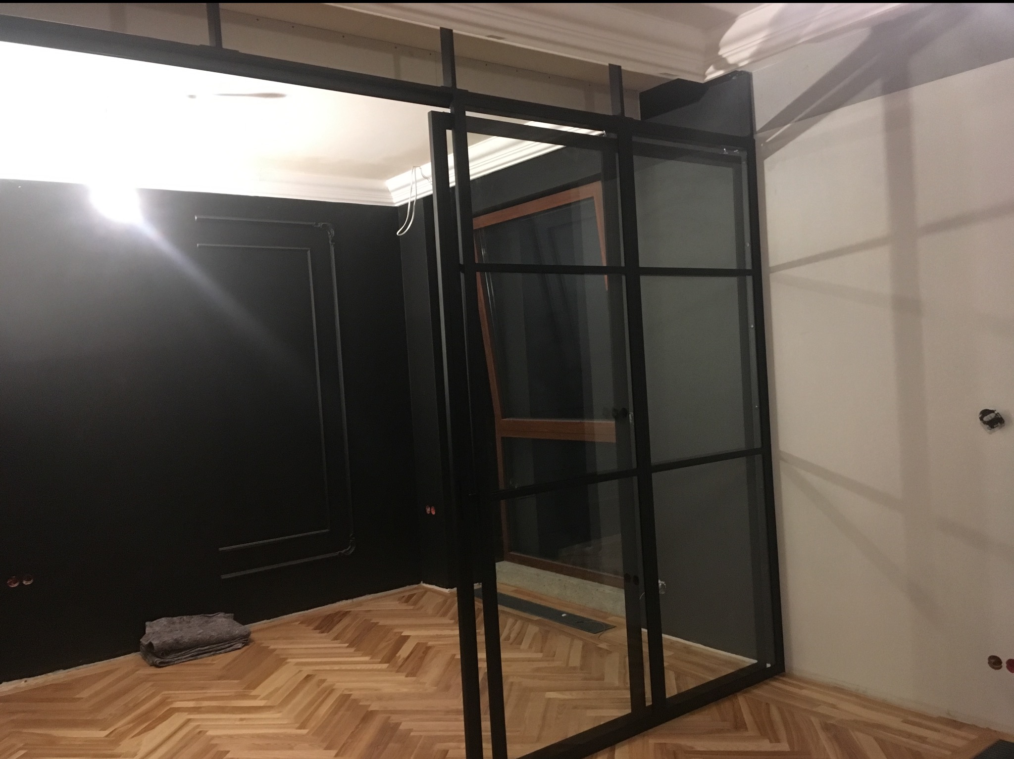 Loftowa, czarna, przeszklona konstrukcja działowa z metalu, oddzielająca pokoje. Widoczny parkiet i fragment czarnej ściany z dekoracyjnymi listwami.
