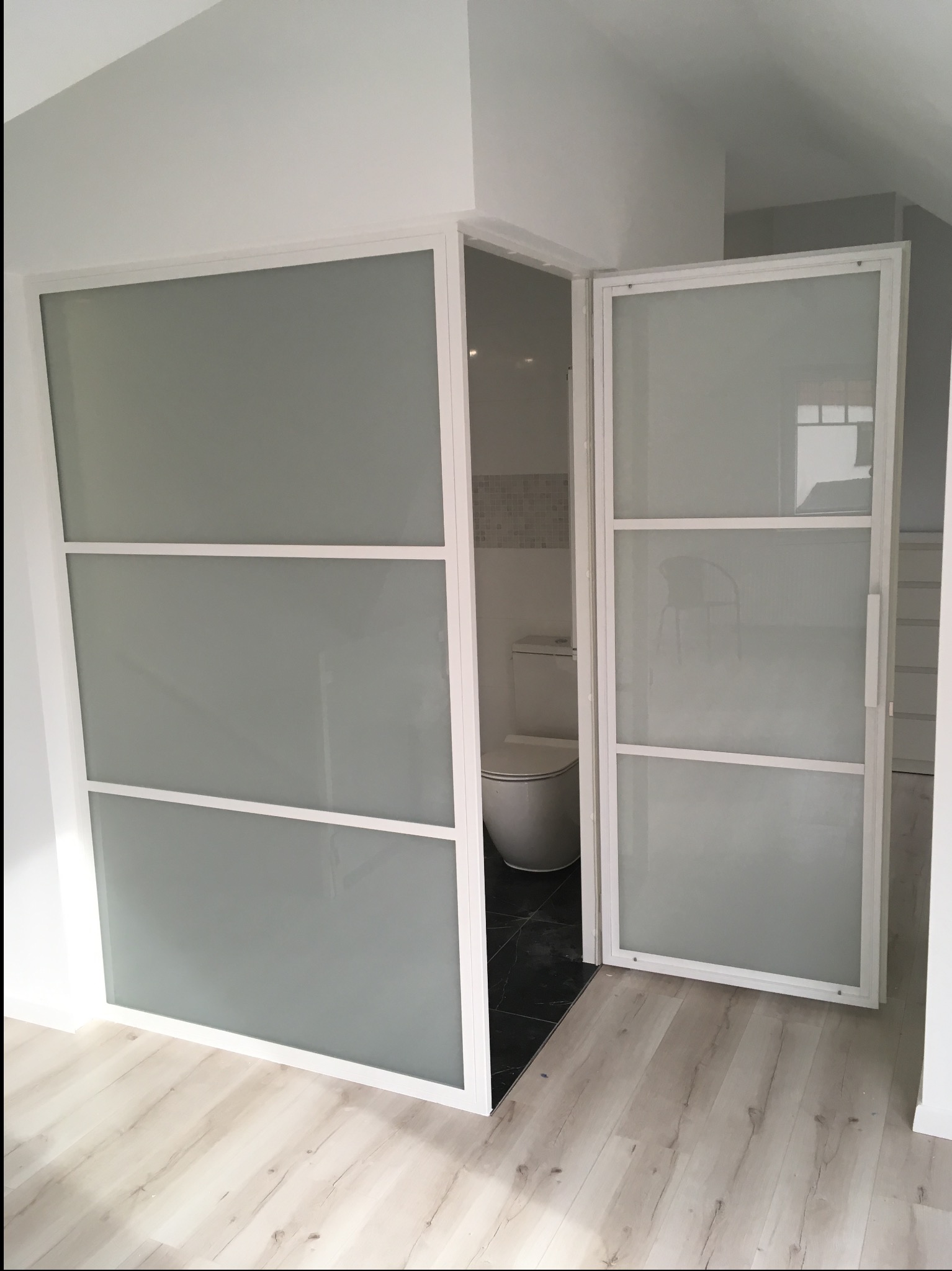 Nowoczesne, białe drzwi szklane z trzema panelami, otwarte do łazienki z czarno-białą mozaiką i toaletą. Widoczne drewniane panele podłogowe.