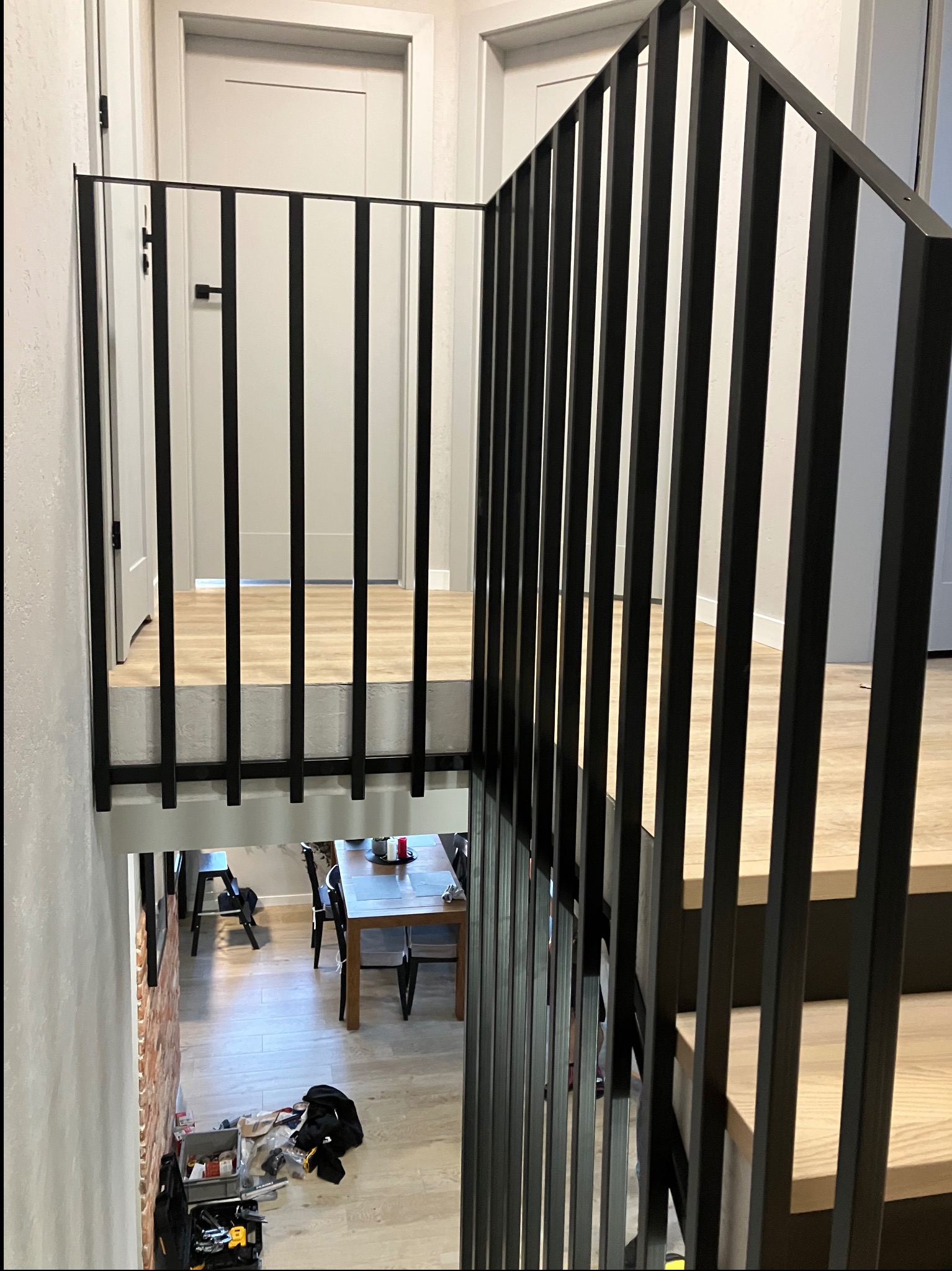 Nowoczesna, czarna balustrada schodowa z pionowymi szczeblami, widok z góry na dół, z widocznymi schodami i fragmentem pomieszczenia z jadalnią w tle.