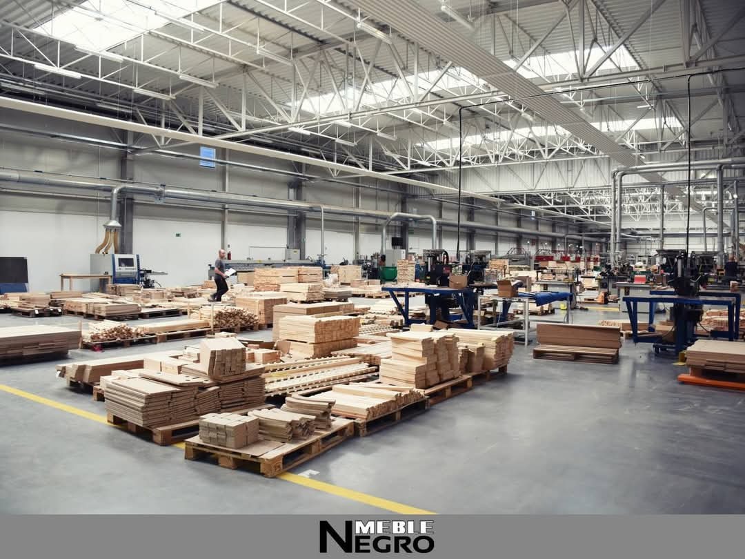 Wnętrze hali produkcyjnej z przygotowanym drewnem na paletach, widoczny pracownik w oddali i maszyny do obróbki drewna.