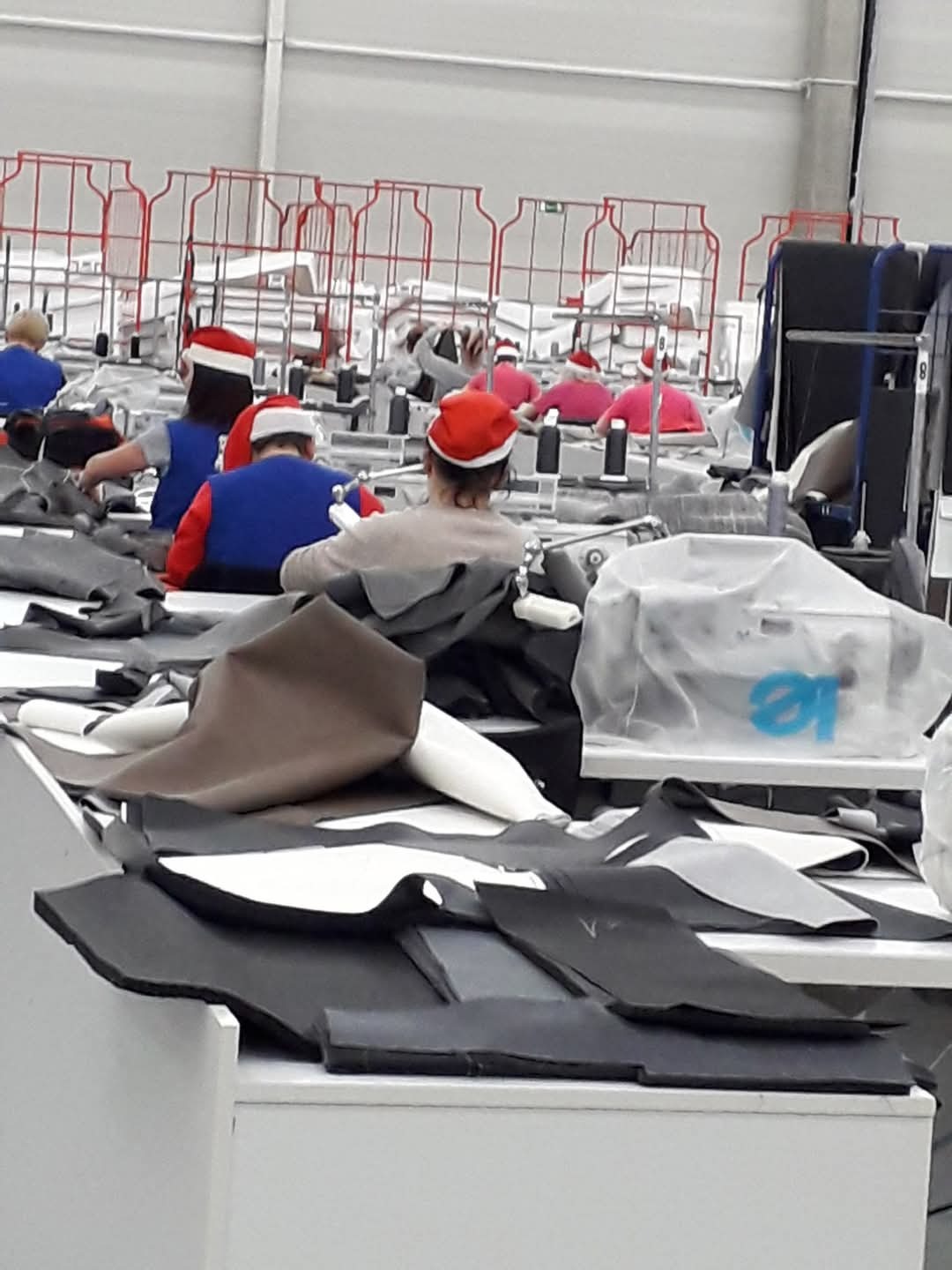 Pracownicy szwalni w czapkach Mikołaja przy stanowiskach pracy, krawędzie materiałów na pierwszym planie, w tle wózki transportowe.