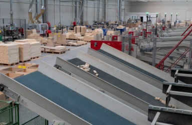 Wnętrze hali produkcyjnej z linią transportującą elementy drewniane, widoczne maszyny i palety z materiałem, oświetlenie górne, perspektywa z góry.