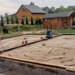 Przygotowany teren pod fundamenty w Gryfinie, z widocznymi ścianami fundamentowymi, zagęszczarką, w tle drewniany dom i domek letniskowy.