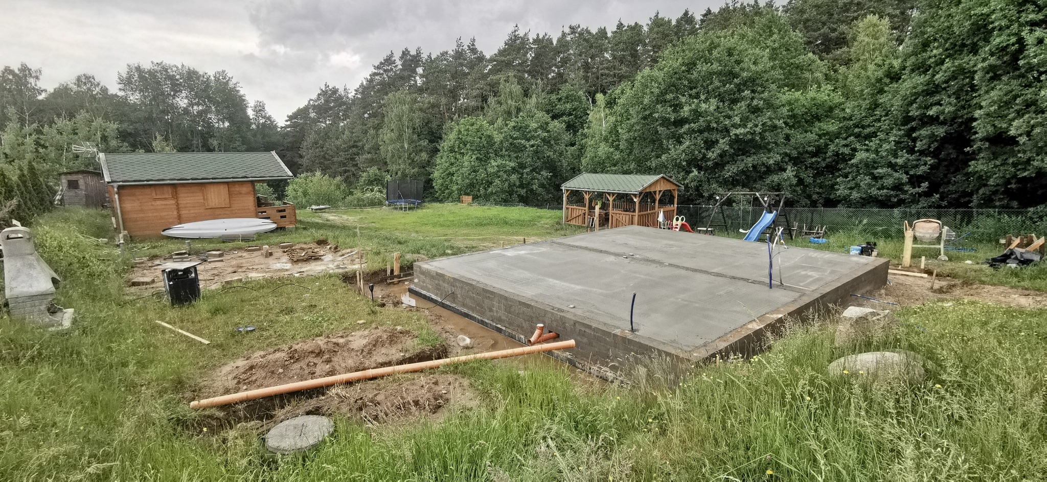 Zalany betonowy fundament z wyprowadzonymi rurami, widoczne przygotowania do dalszych prac budowlanych na działce z domkiem letniskowym i placem zabaw w tle.