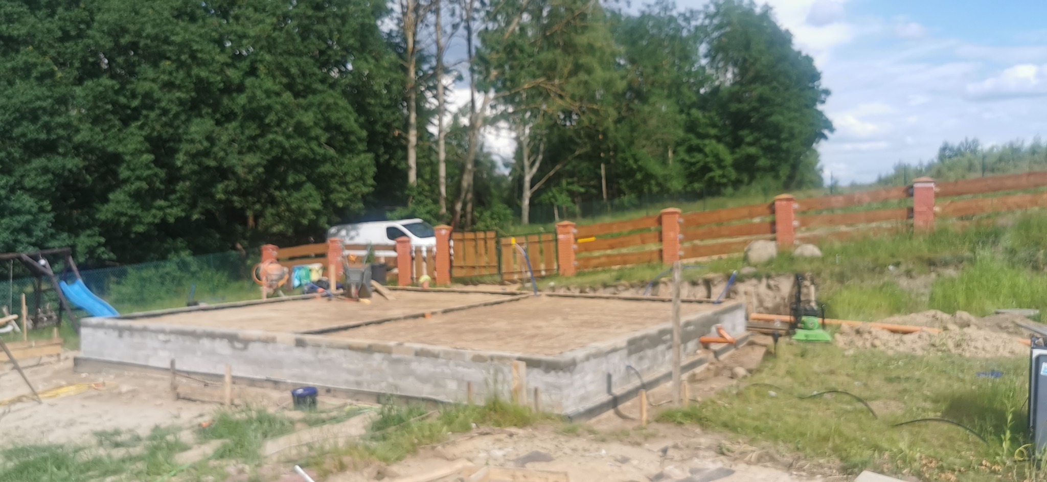 Wylane fundamenty pod budynek jednorodzinny, widoczny szalunek, teren przygotowany pod dalsze prace budowlane, na działce stoi betoniarka i samochód dostawczy.