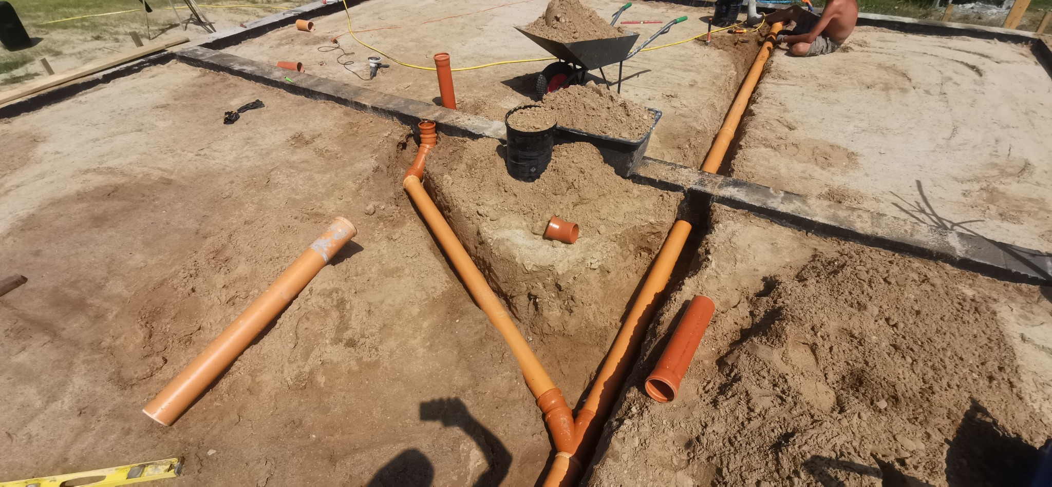 Układanie pomarańczowych rur kanalizacyjnych w wykopie fundamentowym, widoczny fragment szalunku, taczka z ziemią i pracownik przy łączeniu rur.