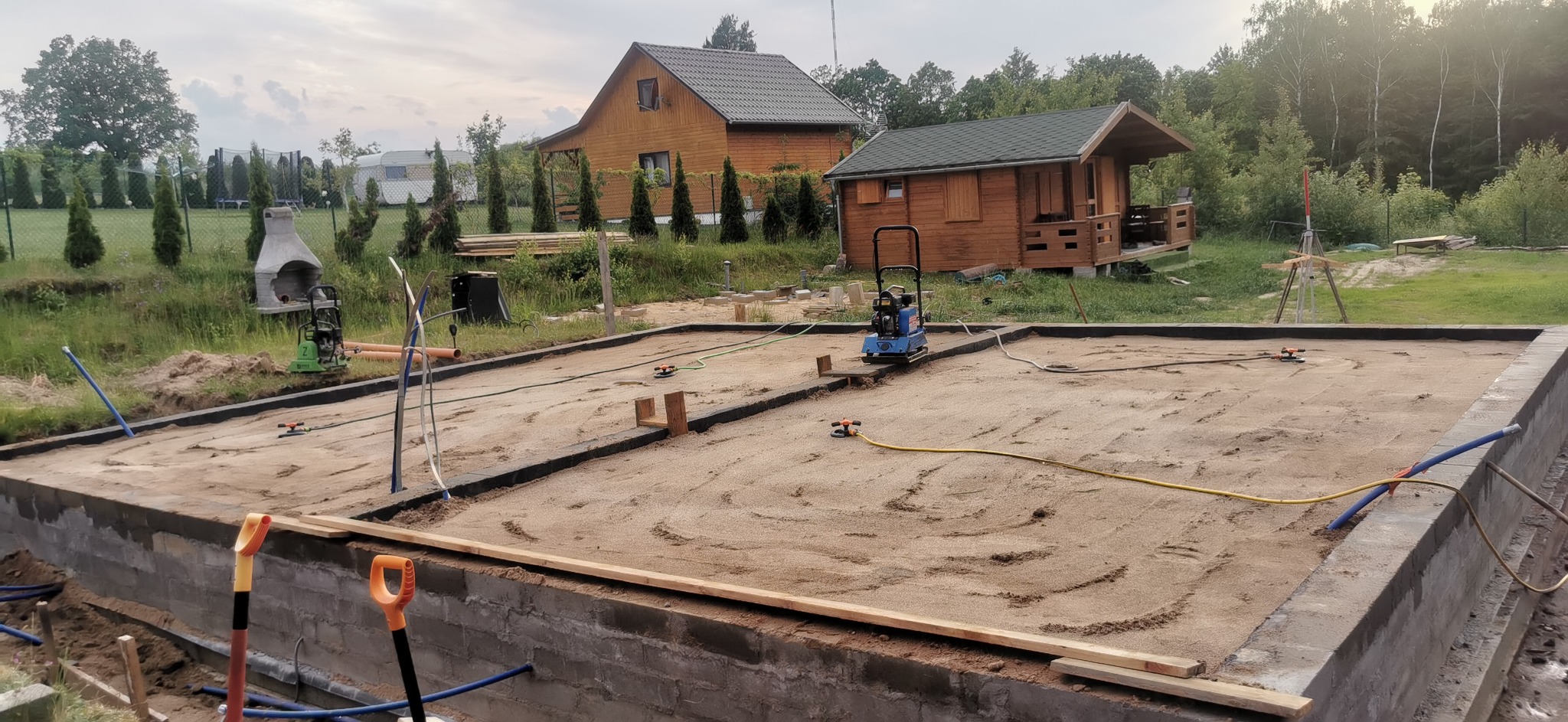 Przygotowany teren pod fundamenty w Gryfinie, z widocznymi ścianami fundamentowymi, zagęszczarką, w tle drewniany dom i domek letniskowy.